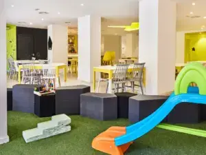 Una cafetería familiar con mesas para adultos, café recién preparado y una zona de juegos infantiles visible al fondo.